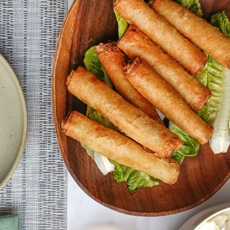 A wooden plate containing crispy lemongrass chicken rolls served with lettuce, alongside a dish of spiced vinegar for dipping.