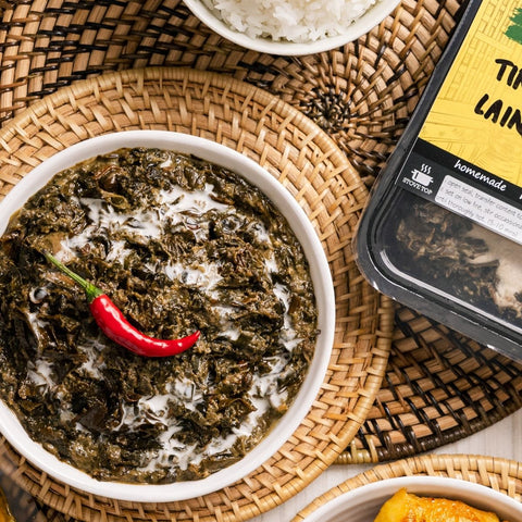 A bowl of tinapa laing, a creamy dish made of callaloo, garnished with a red chili pepper, served on a woven mat. The packaging of the product is also visible showing the product name and branding.