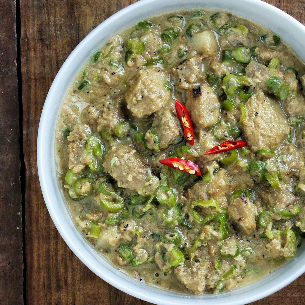 A bowl of Bicol Express, a dish made with tender pork, coconut cream, homemade krill bagoong, and chili peppers, served on a wooden table.