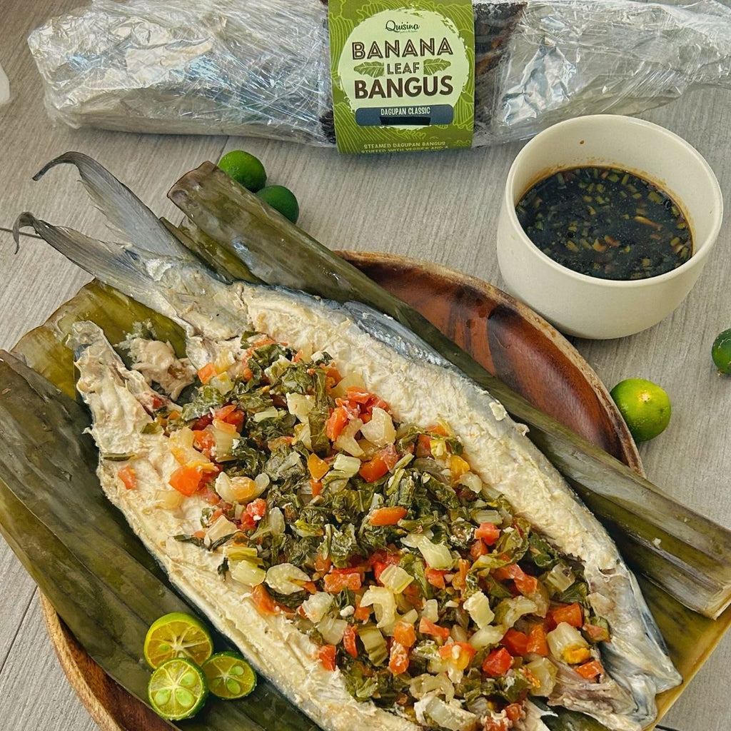A steamed bangus (fish) dish with vegetables on top, served on banana leaves, accompanied by a toyomansi dip and garnished with lime.