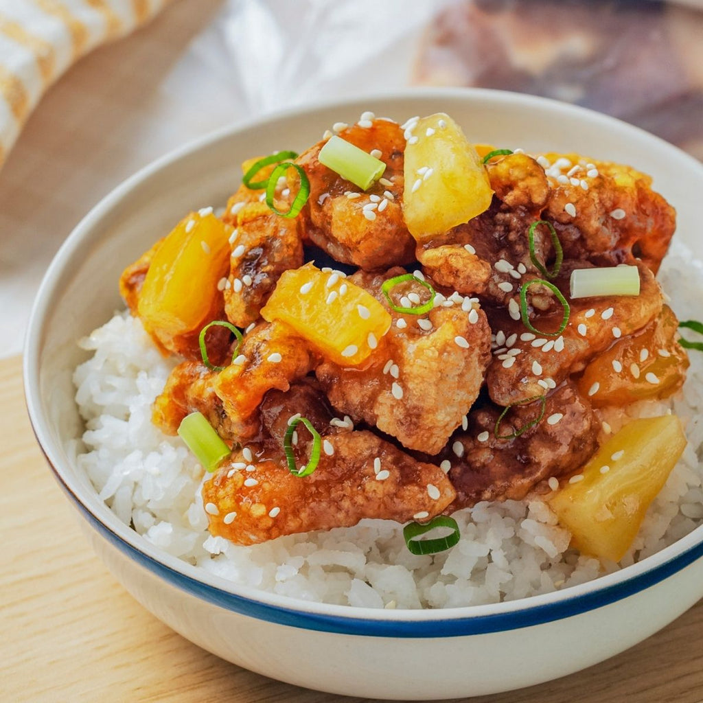 A bowl of sweet and sour pork with pineapple over rice, garnished with sesame seeds and green onions.