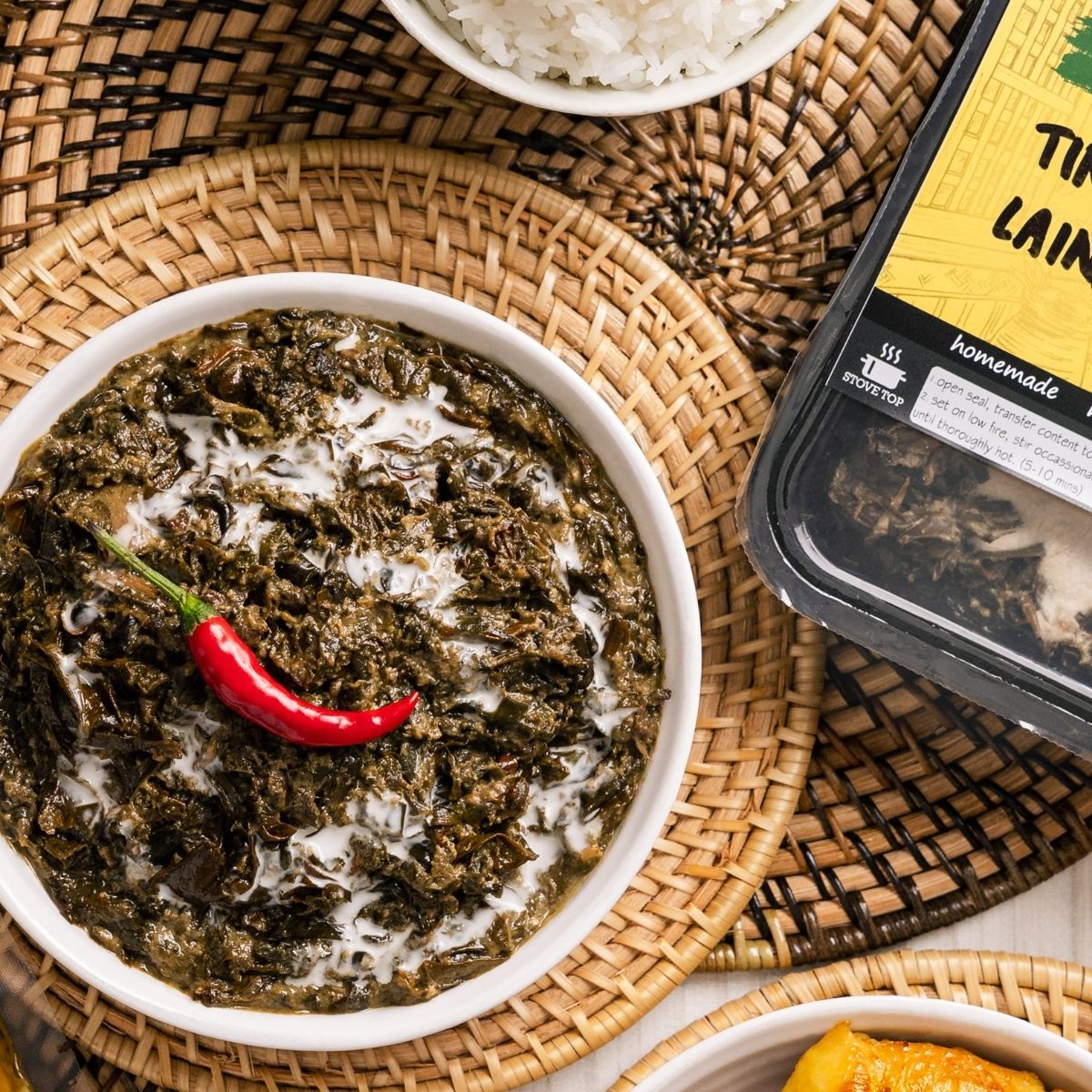 A bowl of tinapa laing, a creamy dish made of callaloo, garnished with a red chili pepper, served on a woven mat. The packaging of the product is also visible showing the product name and branding.